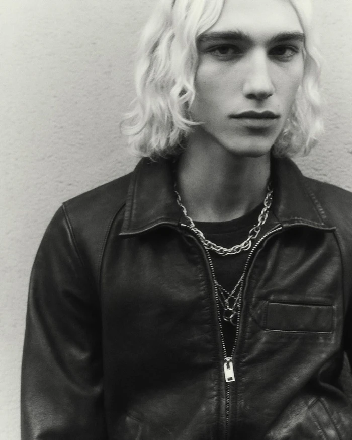 Portrait of a young man with wavy blonde hair wearing a black leather jacket and silver chain necklaces