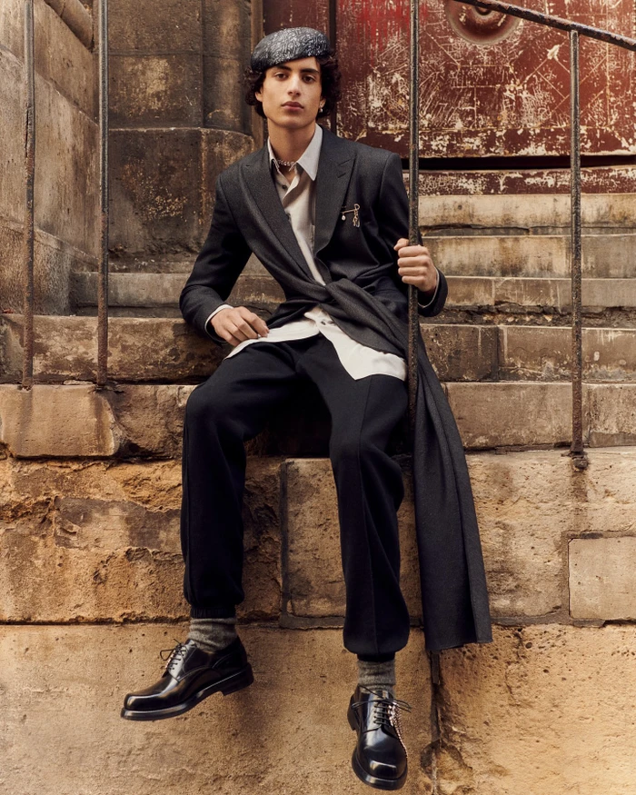 Young male model in dark suit jacket, white shirt, beret, and shiny black shoes seated on stone steps in Paris.