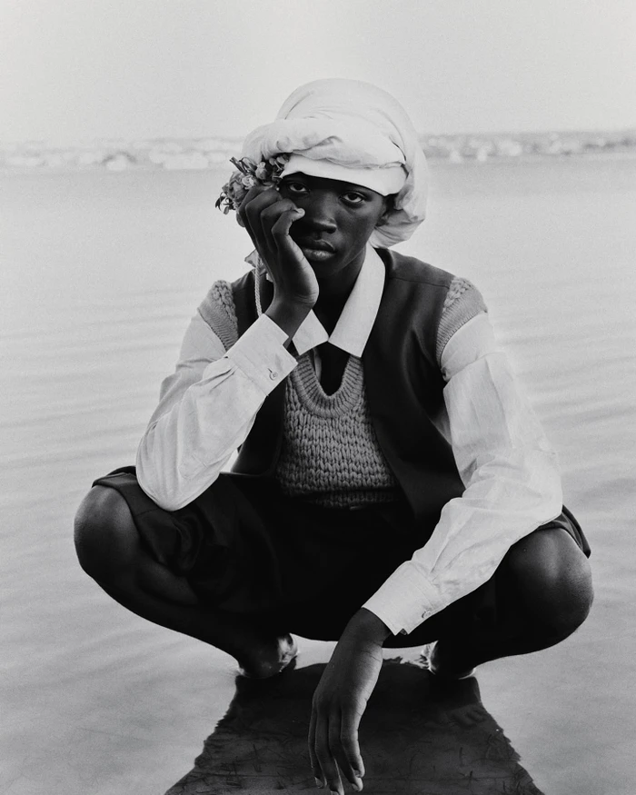 Black man wearing a turban and sweater vest squatting outdoors by the water