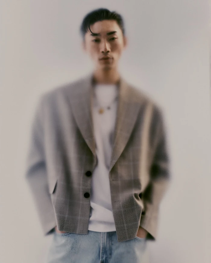 Soft focus portrait of a man wearing a plaid blazer over a white t shirt and blue jeans against a grey background