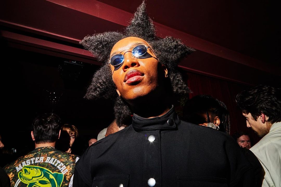 Close-up of an adult with star-shaped hair wearing sunglasses and a dark jacket, surrounded by a crowd in an indoor environment.