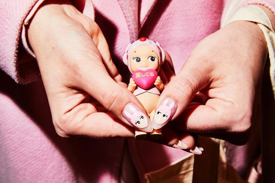Close-up shot of a person holding a Sonny Angel doll with a pink heart in their manicured hands against a pink background.