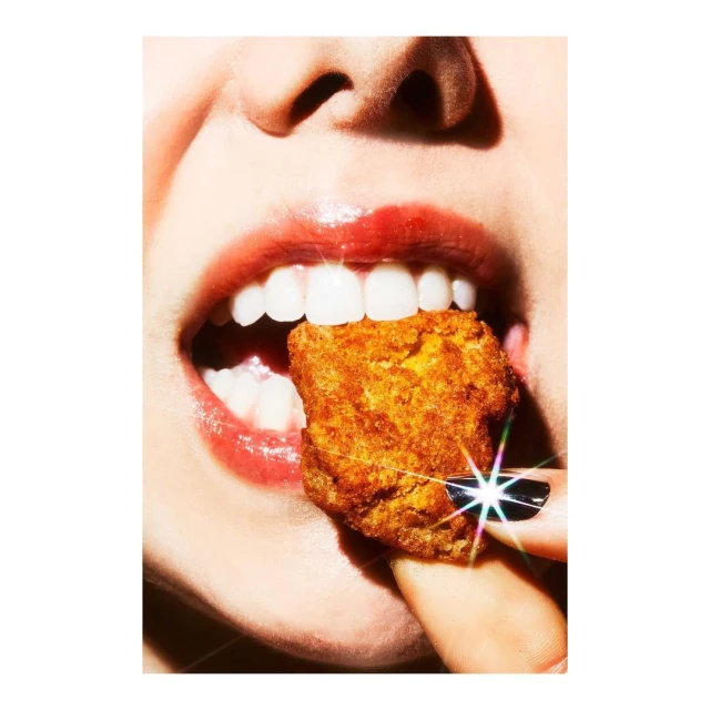 Close-up of a person with red lipstick taking a bite of a fried, golden-brown chicken nugget, set against a bright, clean background.