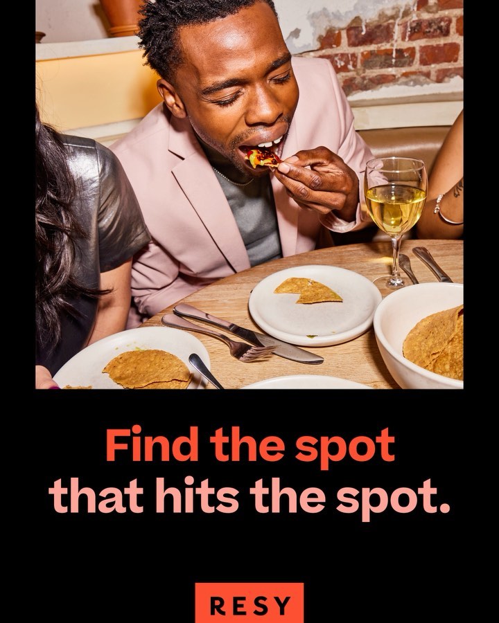 Man in a pink blazer eats food at a restaurant, while two other people sit beside him. Table setting with food and wine.