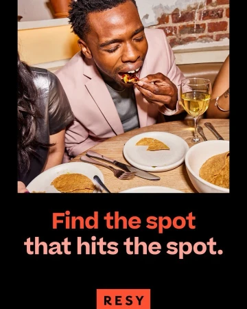 Black man in a pink blazer eating food at a restaurant table with wine and plates, photographed by Amy Lombard.