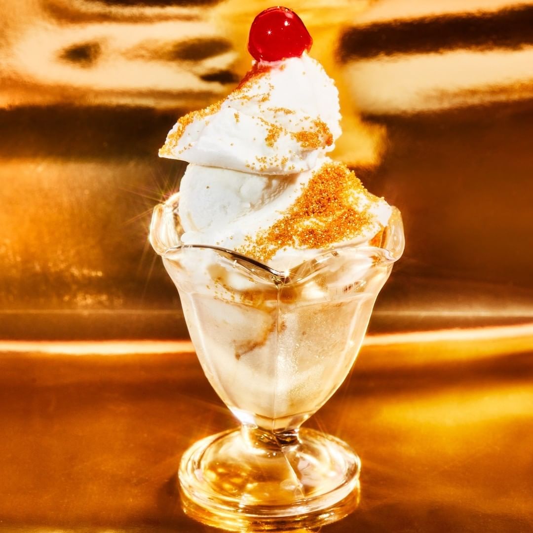 Close-up of an ice cream sundae with whipped cream and a cherry on top in a clear glass bowl on a gold surface.