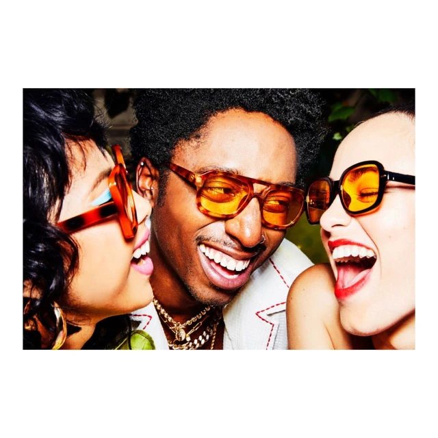 Three joyful people wearing orange-tinted sunglasses are laughing together in a close-up shot that captures their fashionable summer style.