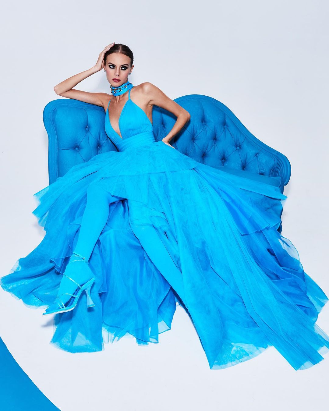 Fashion shoot of a model in a voluminous, bright-blue gown posed on a blue velvet sofa against a bright white background.