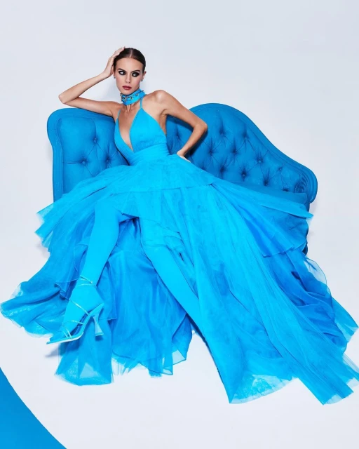 Fashion shoot of a model in a voluminous, bright-blue gown posed on a blue velvet sofa against a bright white background.