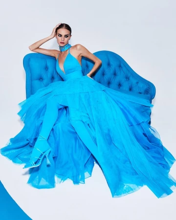Fashion shoot of a model in a voluminous, bright-blue gown posed on a blue velvet sofa against a bright white background.
