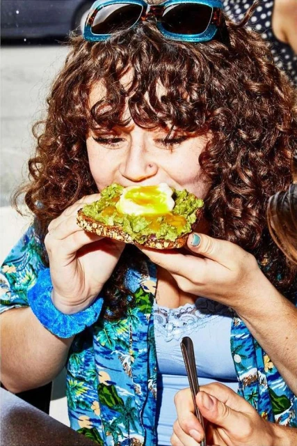 Woman eating avocado toast with egg in an outdoor setting. She wears a tropical shirt, has curly brown hair, and wears blue sunglasses.