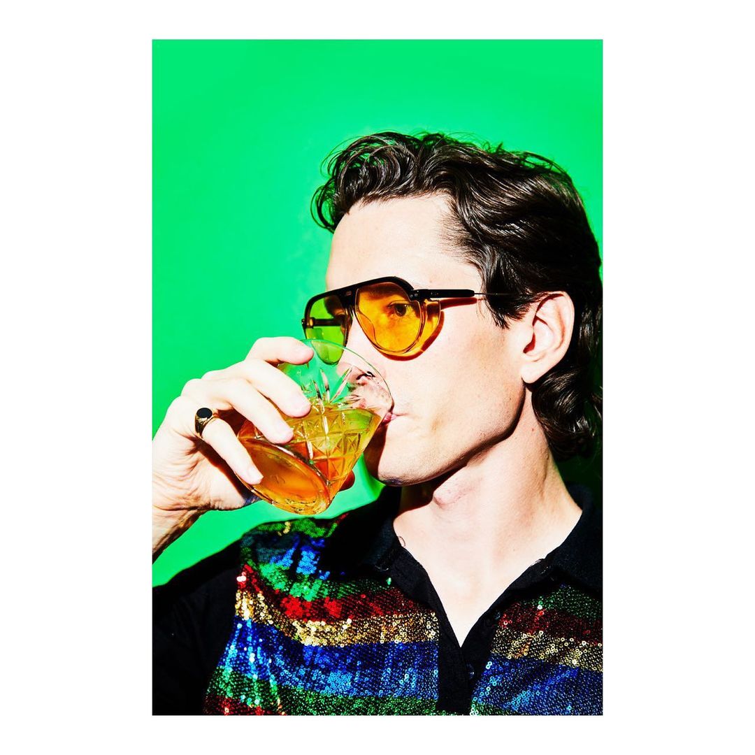 Man in sunglasses drinks from crystal glass against bright green backdrop, wearing sequin shirt and vintage style in a studio shot.