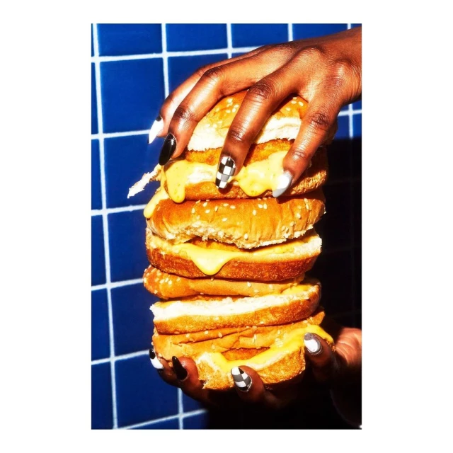 Stacked grilled cheese sandwich held by a dark-skinned adult female hand with checkerboard nail art against a blue tile background.