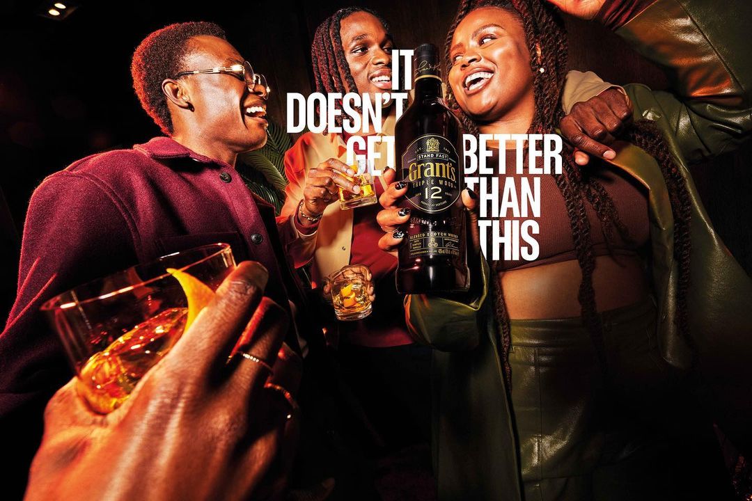 Three young adults are smiling and holding drinks in an indoor bar or event setting, advertising Grants Triple Wood 12 blended Scotch whisky.