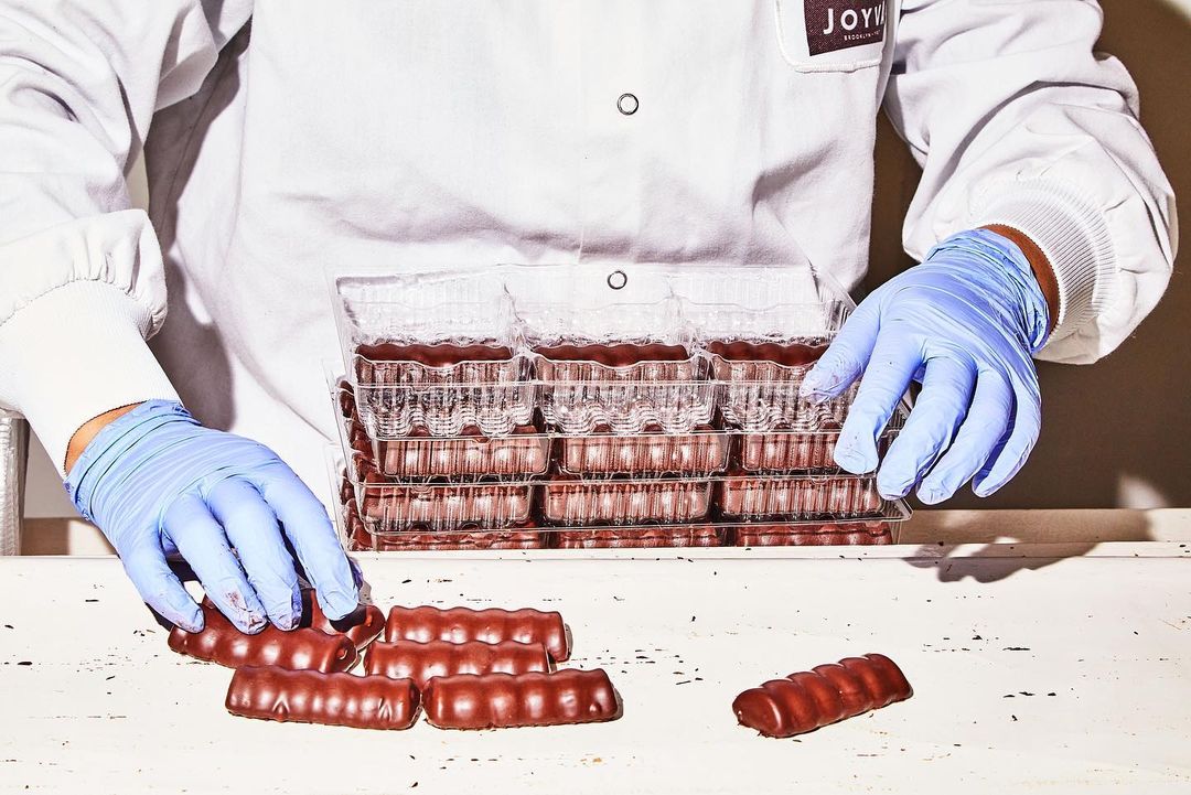 Close-up of a worker in a lab coat and blue gloves sorting trays of chocolate bars on a white table, highlighting food production.