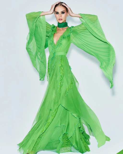 A woman in a flowing lime-green dress with ruffled details and dramatic sleeves poses in a bright, minimalist studio setting.