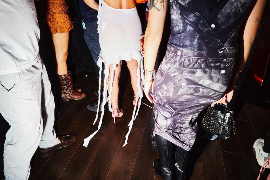Group of people at a party wearing dresses, boots, pants, and carrying a studded purse on a dark wooden floor indoors.