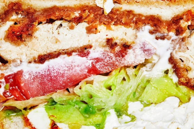 Close-up of a stacked sandwich featuring fried meat, tomato slices, lettuce, and white sauce, showcasing layered textures and vibrant colors.