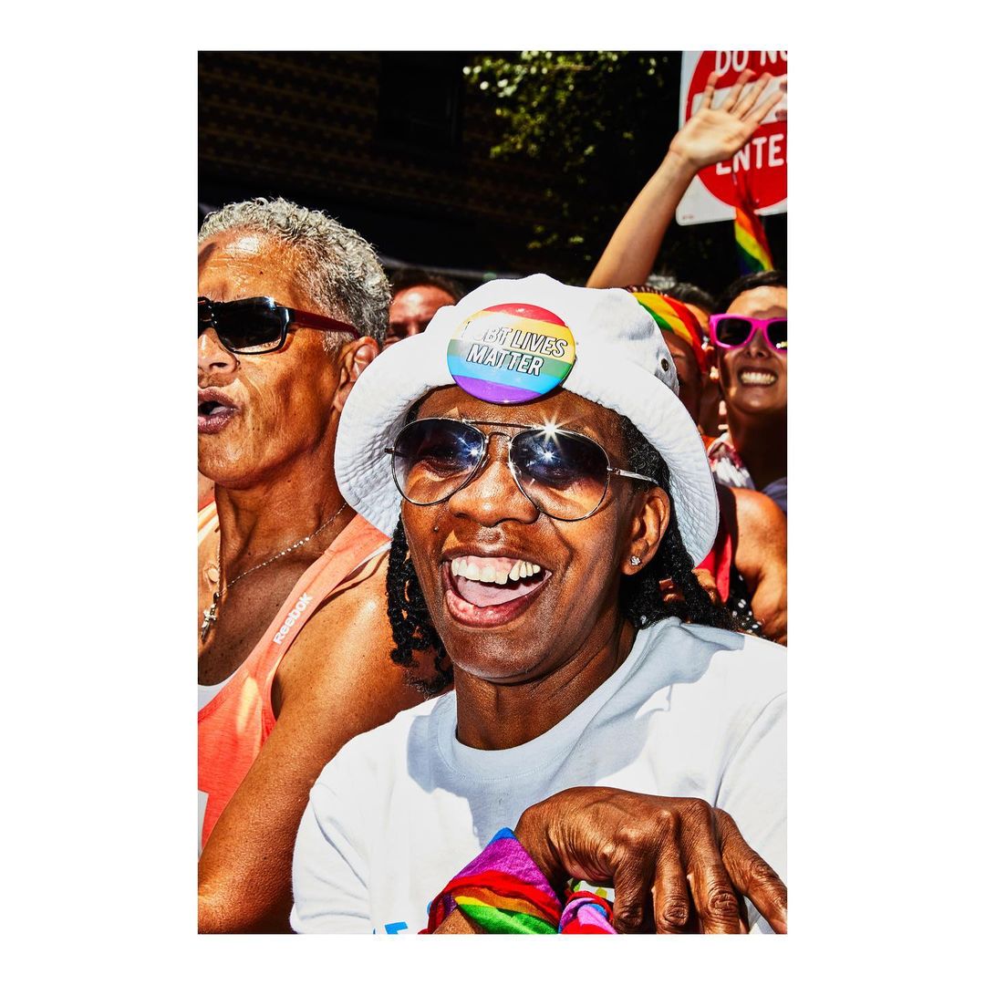 A cheerful crowd celebrates at an outdoor pride parade, with a smiling woman in aviator sunglasses and a 'Gay Lives Matter' button.