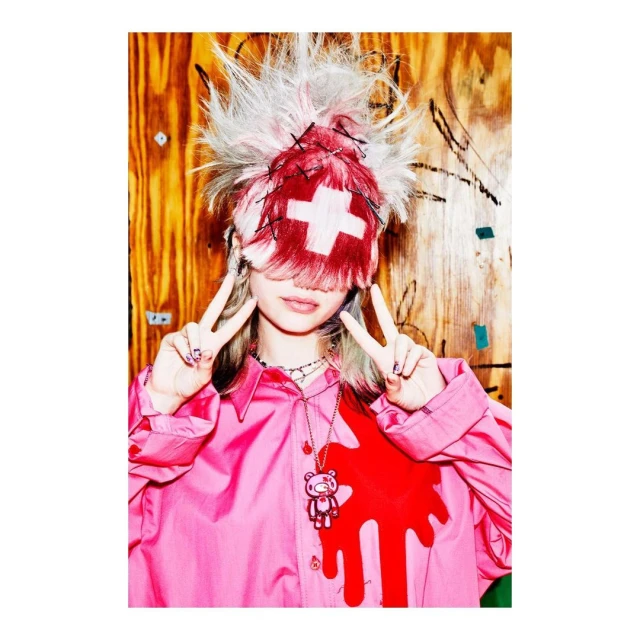 Young woman in pink shirt with unique hat and teddy bear necklace makes peace sign against wooden background with graffiti, indoor shot.