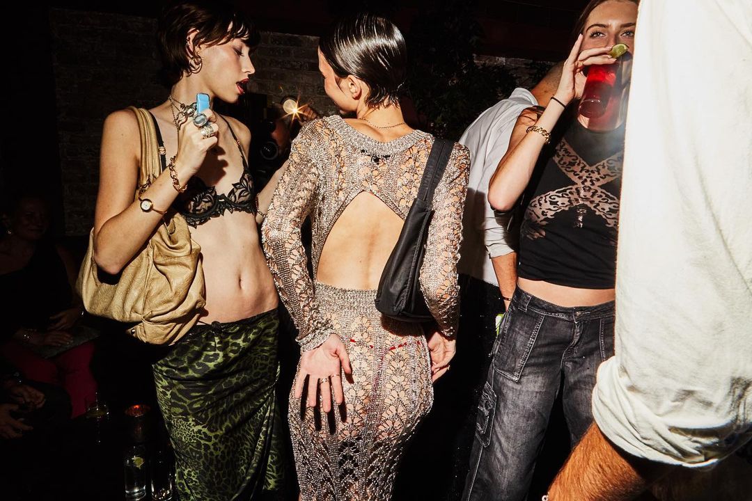 Three women in a dimly lit indoor party environment, wearing fashionable clothing, holding drinks, and socializing under warm lighting.