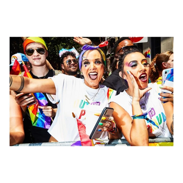 Group of smiling people at an outdoor LGBTQ+ pride parade. People are holding cell phones, wearing rainbow-themed clothing, and bright makeup.