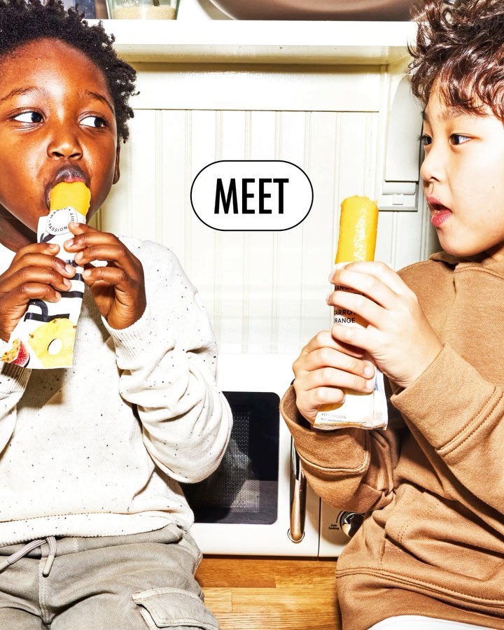 Two young children, an African-American boy and an Asian boy, sit near a microwave and eat orange ice popsicles indoors.