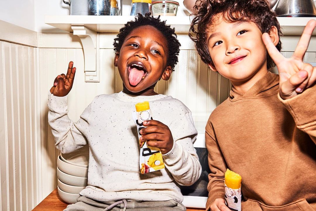 Two smiling children indoors, one sticking tongue out, the other making a peace sign while holding ice pops in a kitchen.