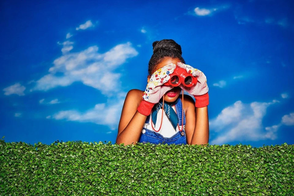 A stylish Black woman peers over a green hedge with red binoculars against a bright blue sky with fluffy white clouds background.