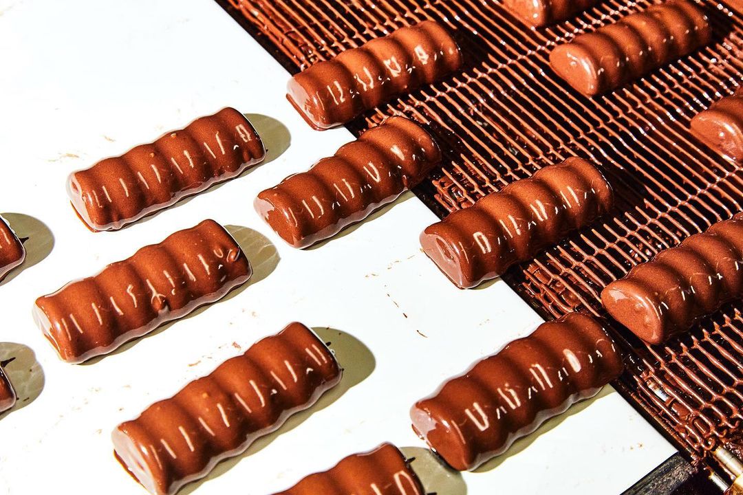 Rows of glossy chocolate-covered candy bars on a production line, featuring a brown conveyor belt and bright, even studio lighting.