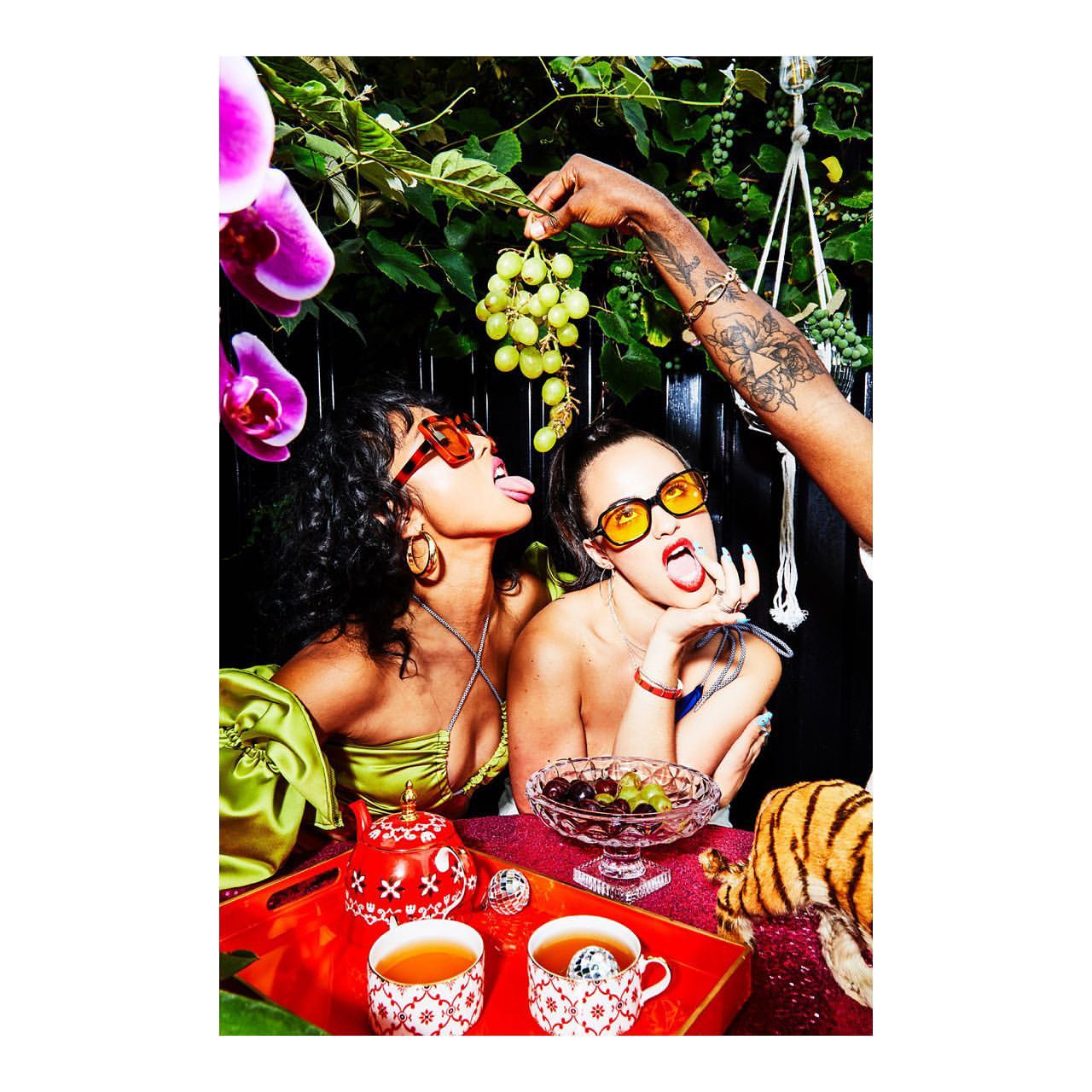 Two women with sunglasses enjoy green grapes at a table with tea cups, orchids, and greenery in an indoor setting.