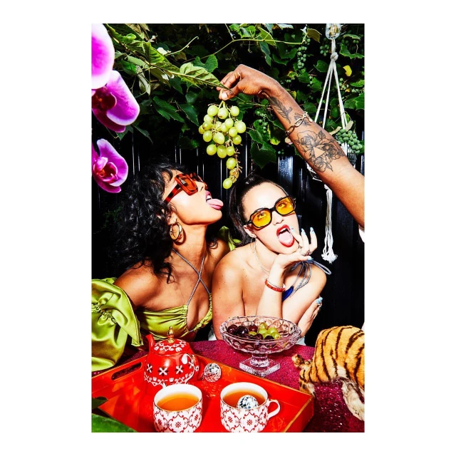 Two women with sunglasses enjoy green grapes at a table with tea cups, orchids, and greenery in an indoor setting.