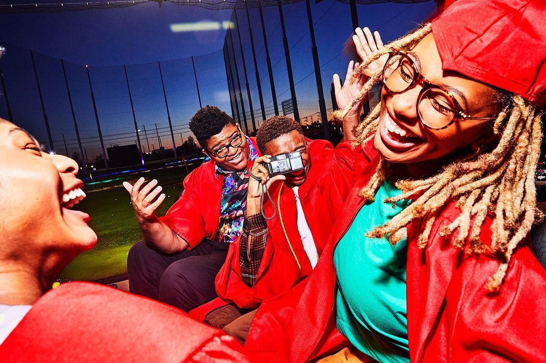 A joyous group of young adults celebrate graduation at a Topgolf-like venue, dressed in red gowns, laughing, and taking photos.