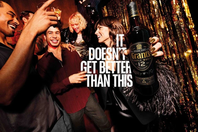 A group of young adults are together indoors at a social gathering with a bottle of Grant's Triple Wood 12-year-old Scotch whisky.