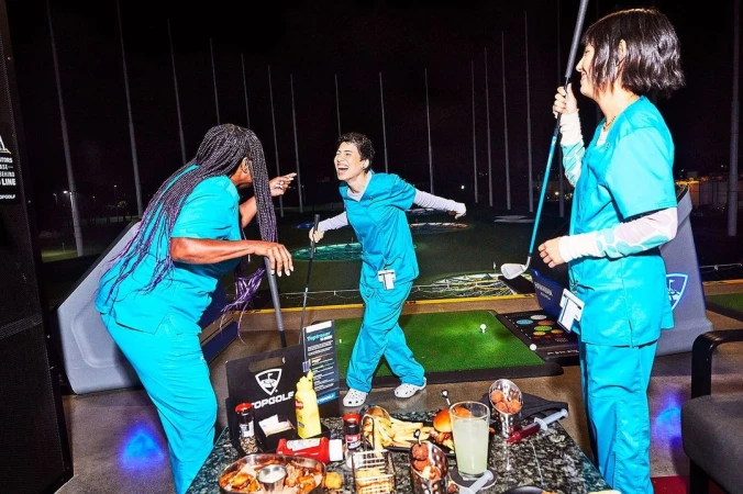 Three women in scrubs laugh and play golf at night at a Topgolf location, with food and drinks on a table in the foreground.