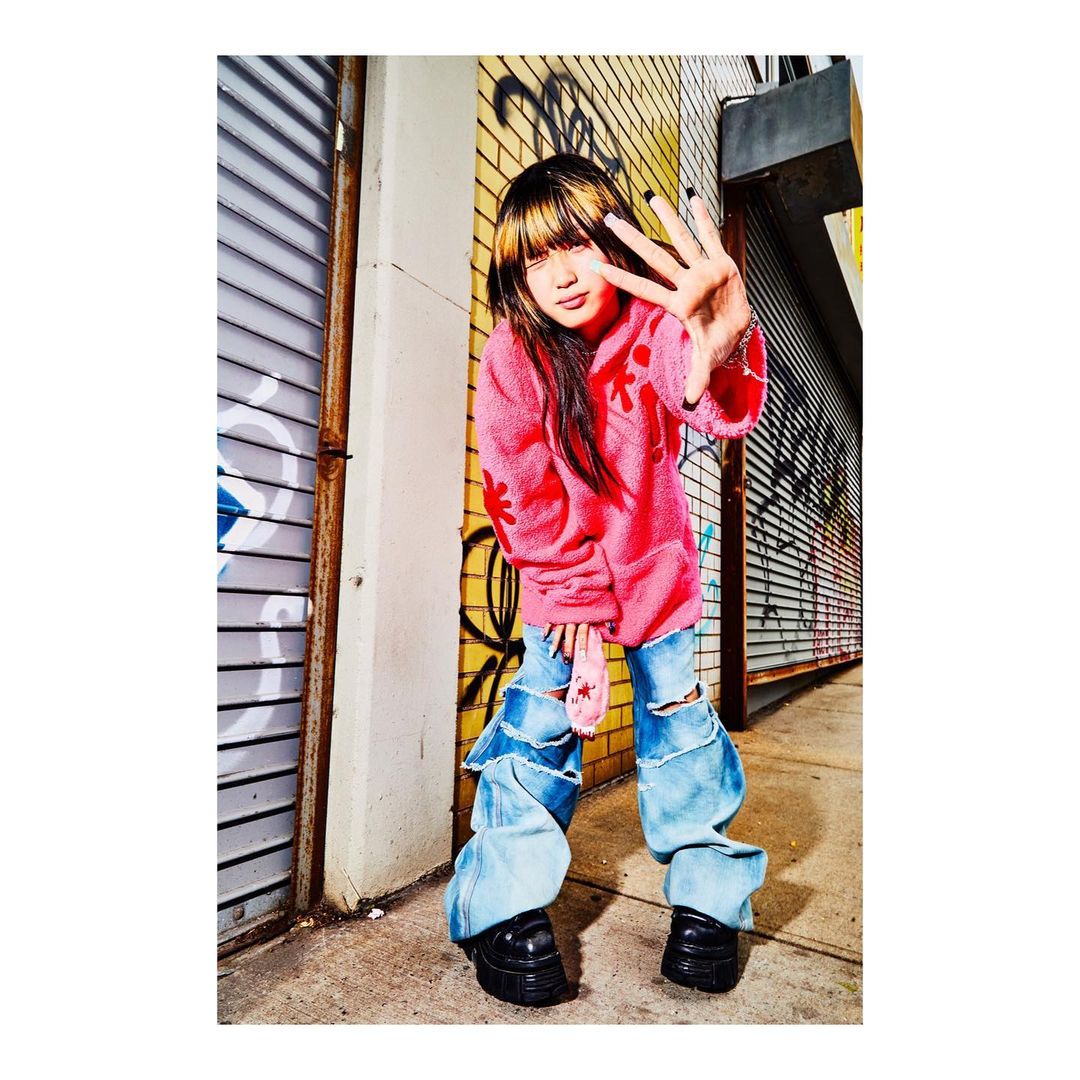 Teenage female with bangs poses in baggy denim jeans and a pink top against graffiti urban wall, holding up four fingers.