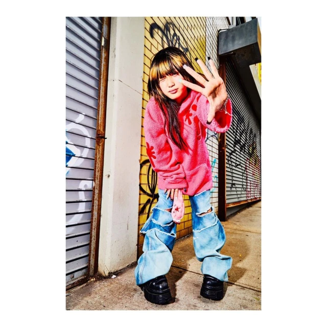 Teenage female with bangs poses in baggy denim jeans and a pink top against graffiti urban wall, holding up four fingers.