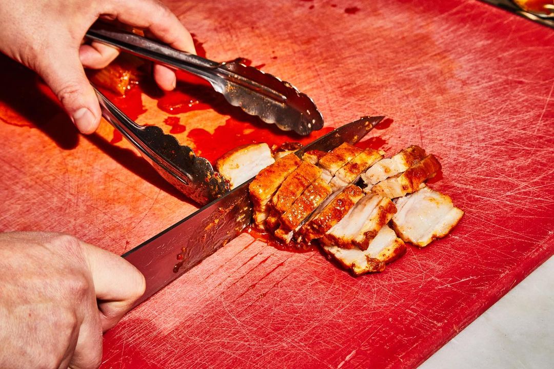 Close-up shot of hands using a knife and tongs to slice cooked pork belly on a red cutting board, showcasing culinary food preparation.