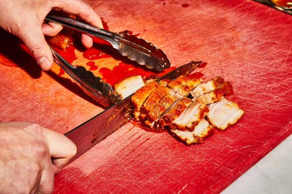 Close-up shot of hands using a knife and tongs to slice cooked pork belly on a red cutting board, showcasing culinary food preparation.