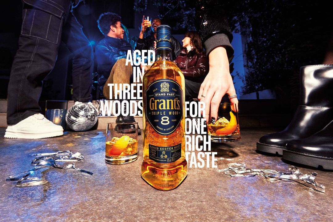 Bottle of Grant's Triple Wood 8 scotch whiskey with cocktails, disco ball, and people in a casual gathering at night.