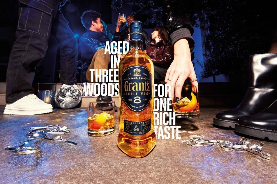 Bottle of Grant's Triple Wood 8 scotch whiskey with cocktails, disco ball, and people in a casual gathering at night.