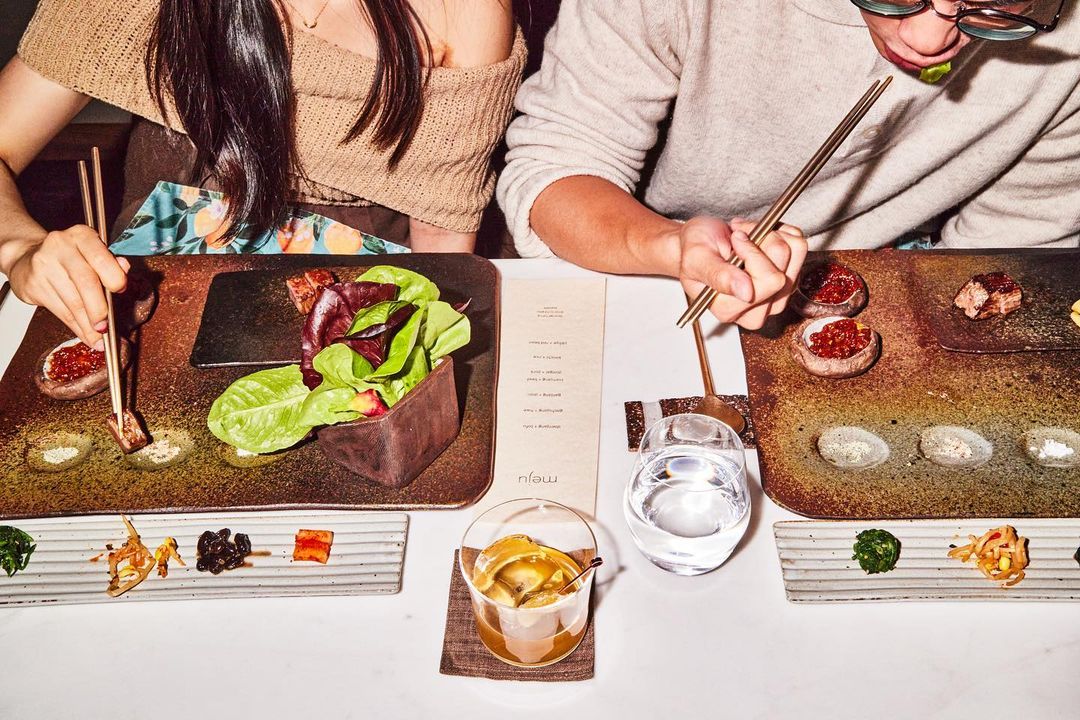 Overhead shot of a couple at a restaurant, enjoying Korean barbeque with various side dishes, salads, and drinks on a bright table.