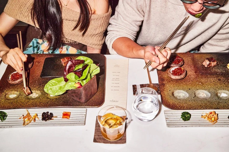 Overhead shot of a couple at a restaurant, enjoying Korean barbeque with various side dishes, salads, and drinks on a bright table.