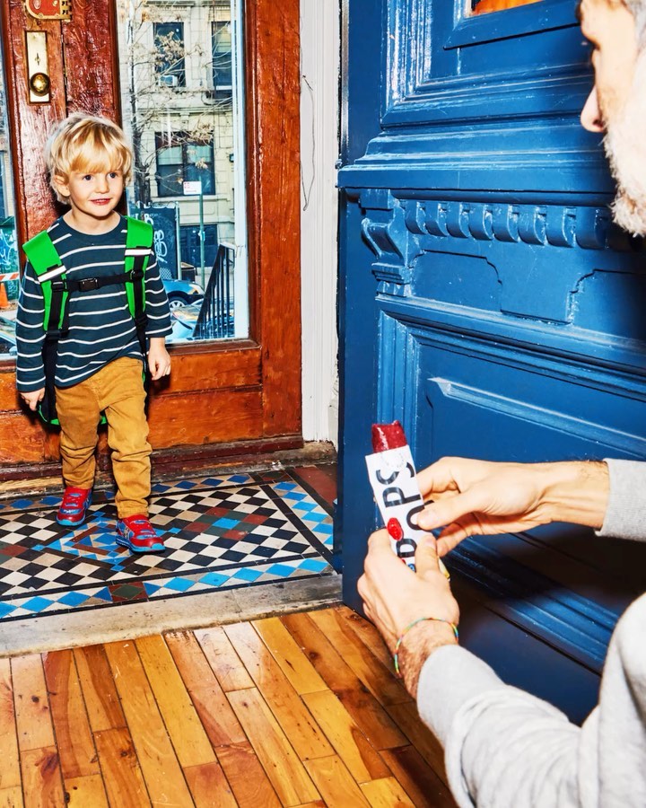 Young boy with blond hair and a green backpack stands in doorway as person hands him a popsicle in front of a blue door.