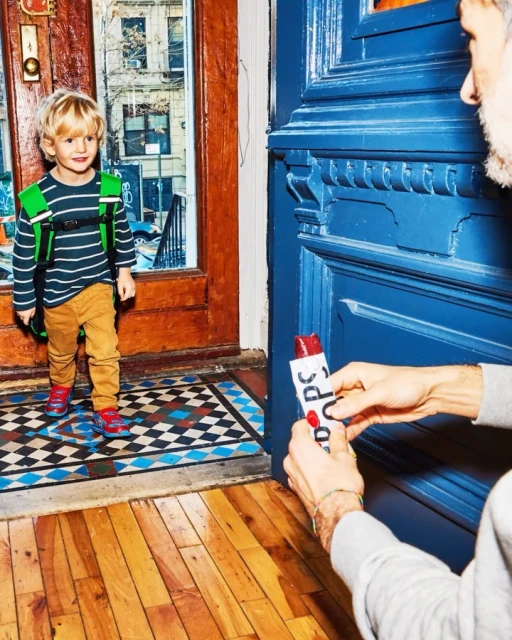 Young boy with blond hair and a green backpack stands in doorway as person hands him a popsicle in front of a blue door.