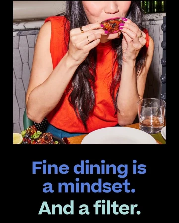 Woman in red sleeveless shirt eats a chicken wing indoors at a table with drinks and salad. Marble wall paneling in the background.