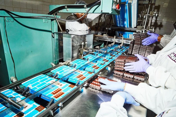 Food factory workers in lab coats packaging chocolate candy bars on an assembly line with automated machinery and conveyor belts.