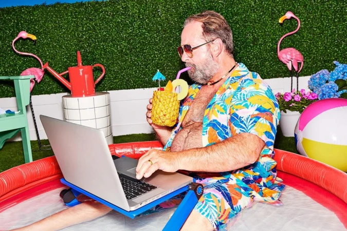 Bearded man in a tropical shirt sits in a kiddie pool, working on a laptop, and sipping a pineapple drink outdoors.