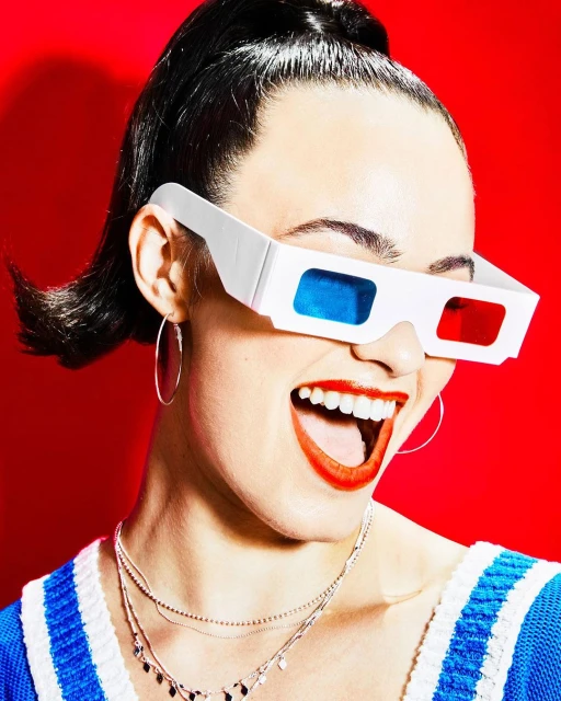 Close-up of a smiling woman wearing white 3D glasses with red and blue lenses, bright red lipstick, and a blue and white sweater.