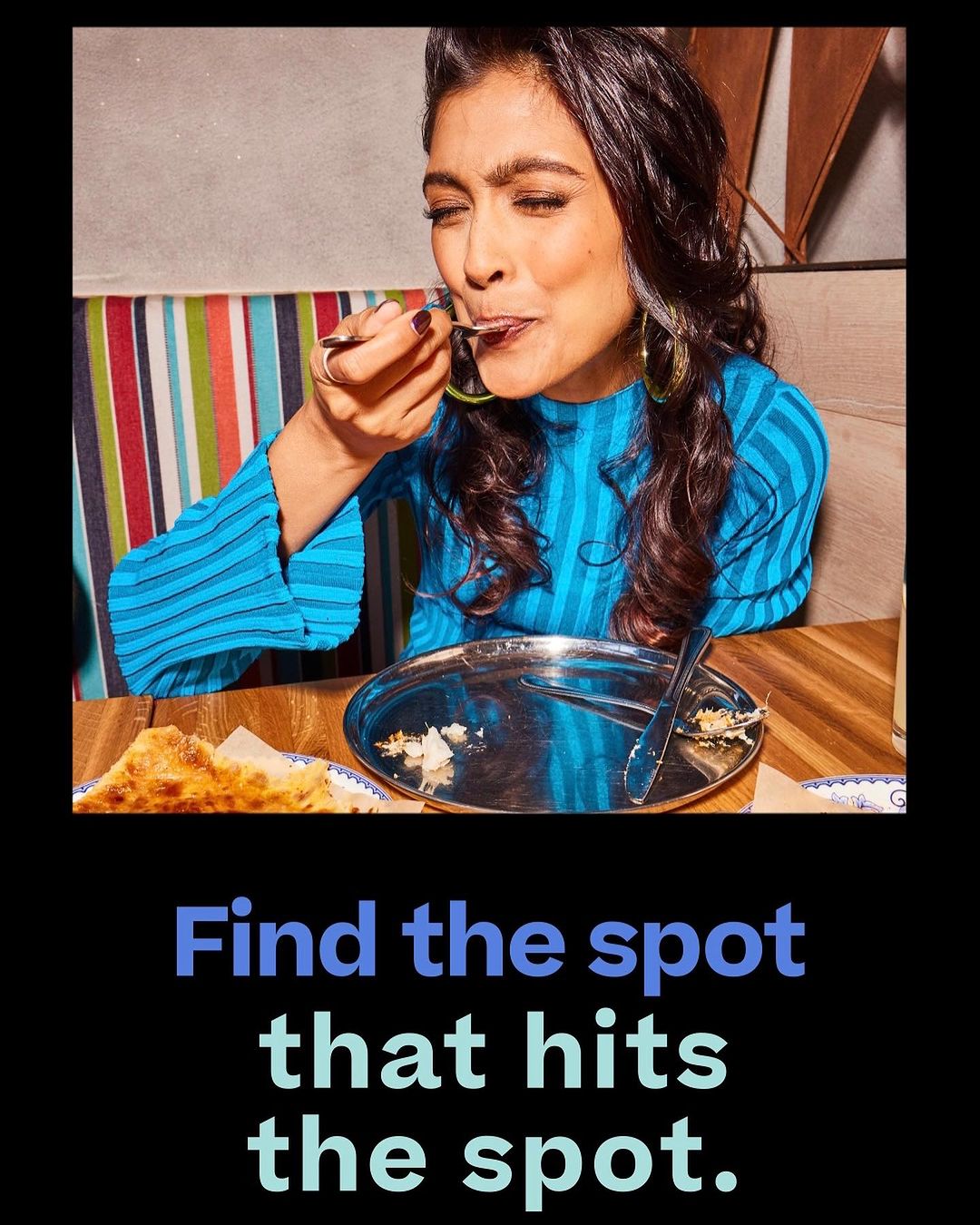 Woman with dark hair in a blue top enjoys a dessert with a spoon. Table holds a metal plate and fork. Backdrop has striped fabric.